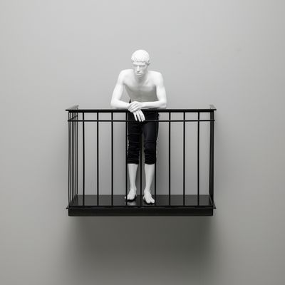 minature white figurine wearing black trousers on metal balcony, mounted on a pale grey wall
