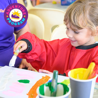 A child in a red smock is painting with a brush on paper, surrounded by cups of colorful paint. A round "Adventure Program" logo is seen nearby.