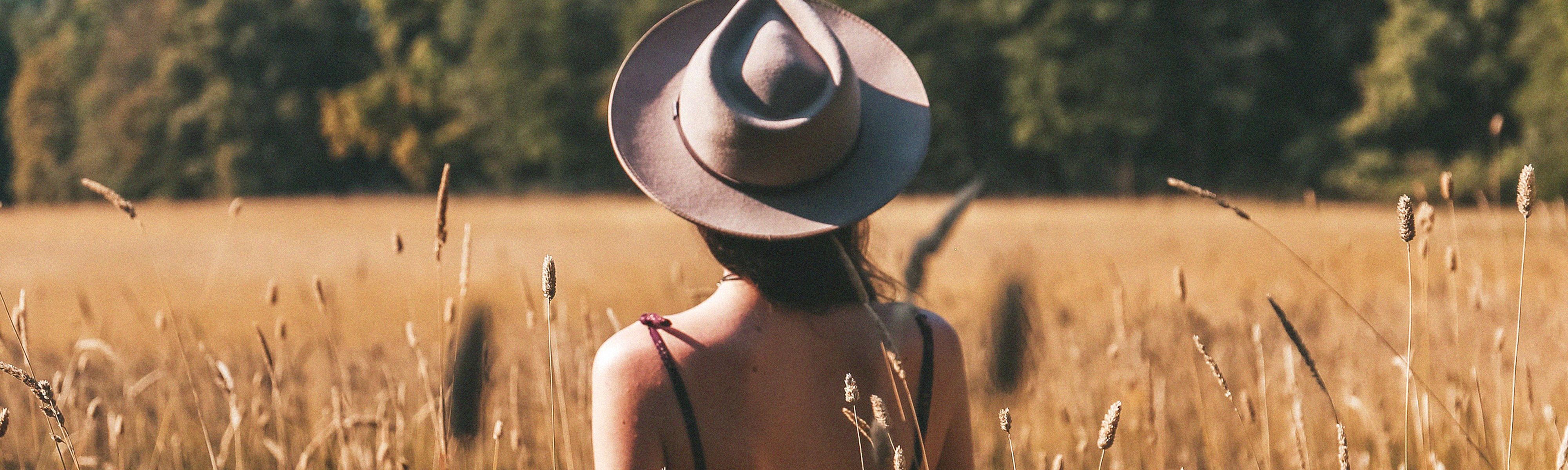 back of womens head wearing a tan hat walking through grass field