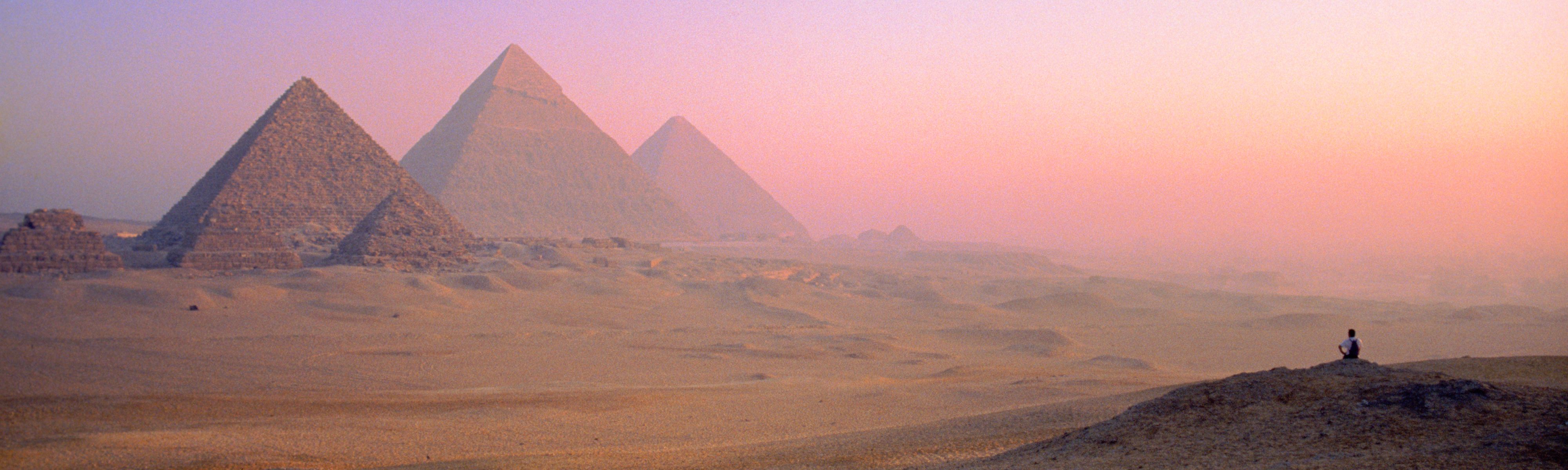 Man sitting on hill looking at Great Pyramids of Giza at sunset