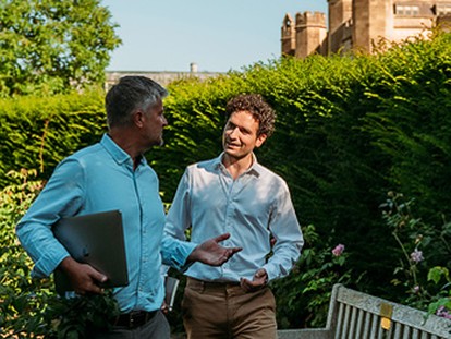 Two people walk in the garden, tall hedges overhead, while having a conversation in the sun