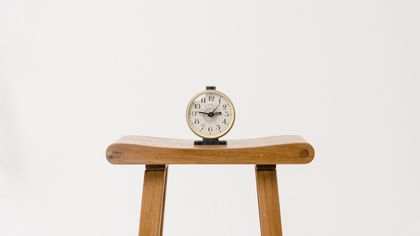 An image of a small alarm clock on a wooden stool against a white background.
