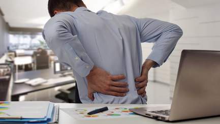 Mann sitzt mit Rückenschmerzen am Schreibtisch und versucht sie zu lindern