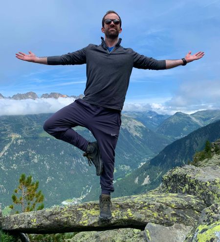 A hiker doing a yoga pose with mountains and the blue sky behind him.