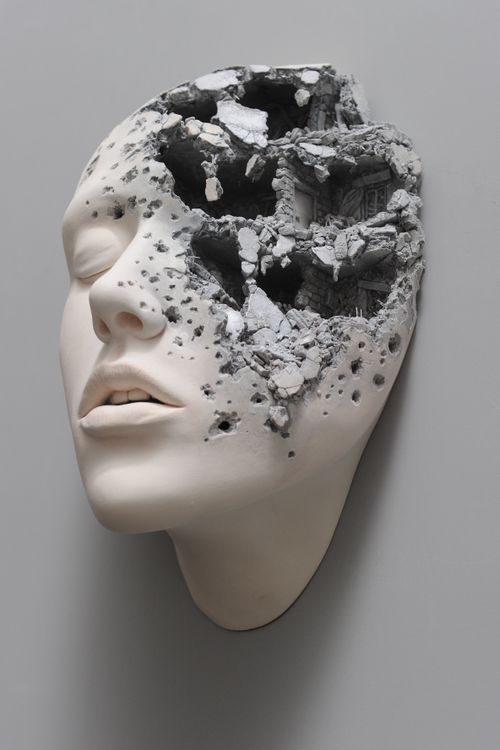 a white sculpted head emerging from a wall with half of the face crumbling inwards like a house demolition, revealing tiny brick walls inside the head