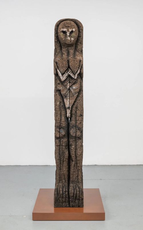 long vertical block with bodily details of a human figure carved into it, placed on a small pedestal