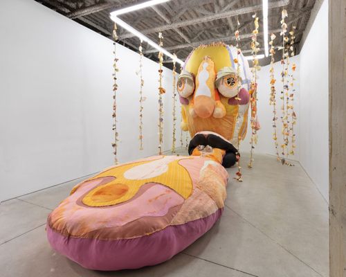 installation view of a giant yellow head with bulging eyes and a long nose sewed on, with garlands of flowers falling from the ceiling around it and an enormous tongue sticking out and trailing along the floor