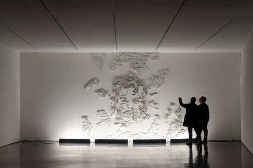 installation view of a dark room with a white wall with a male face illuminated and emerging from it