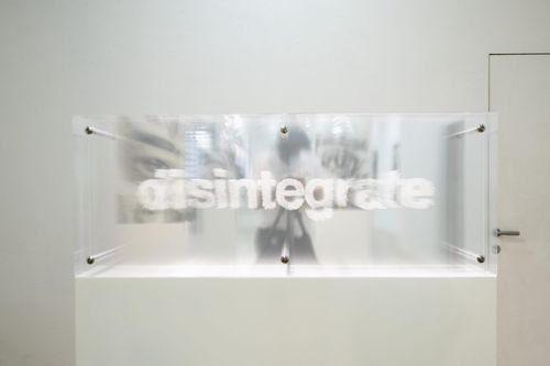 installation view of white room with a glass rectangle that says 'disintegrate' in white writing, placed on a white plinth