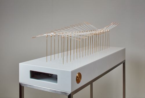 small sculpture sitting on table surface, made up of dozens of small vertical poles set in two lines, connected to the opposite pole by a curved horizontal pole that sits on top, creating a tunnel through the centre