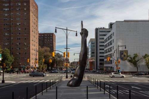A street in New York City with a pedestrian space in the middle of the road, where a large sculpture of a forearm and a finger pointing upwards emerge from the ground 