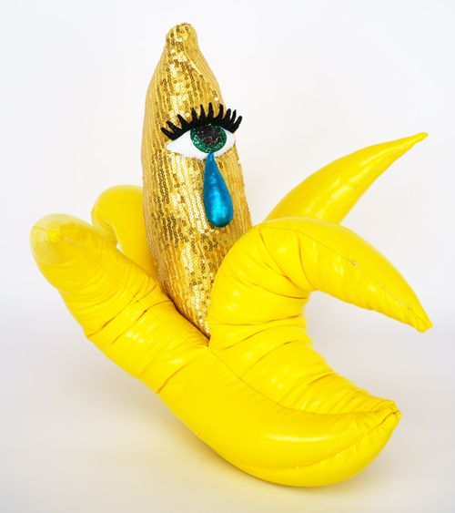 A banana half-peeled and covered in yellow sequins, with an eye sewn onto it which sheds a tear