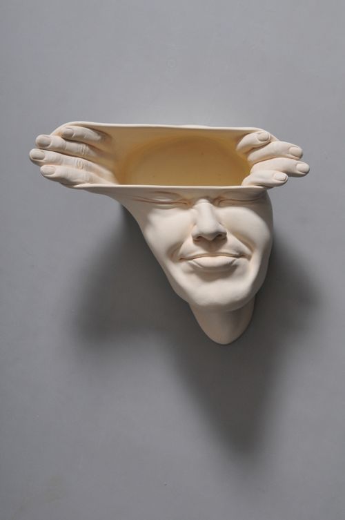 a three-dimensional sculpted head with two hands that reach out of the top of the head and stretch the skin horizontally