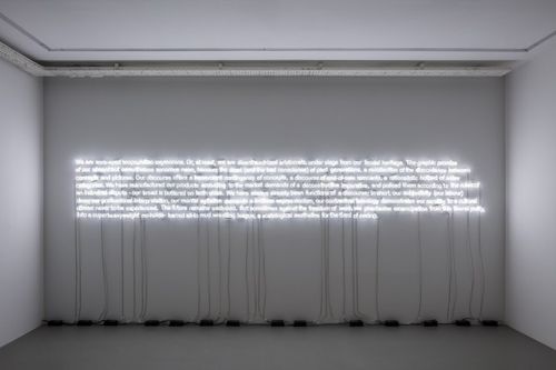 Neon lights illuminating a paragraph of text that hangs centrally in a white room