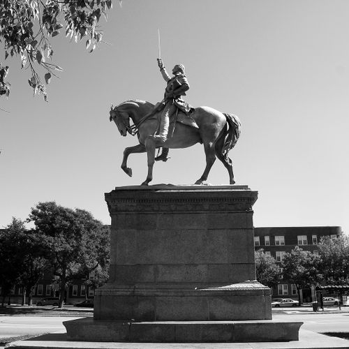 Mounted rigidly atop a powerful horse striding forward, George Washington is shown attired in full military dress, with tricorn hat held in the crook of his left arm and sword raised high above his head in his right hand. The horse and rider, raised high above our heads on a large pedestal, face south in the direction of Washington Park from an island on Dr. Martin Luther King Boulevard.