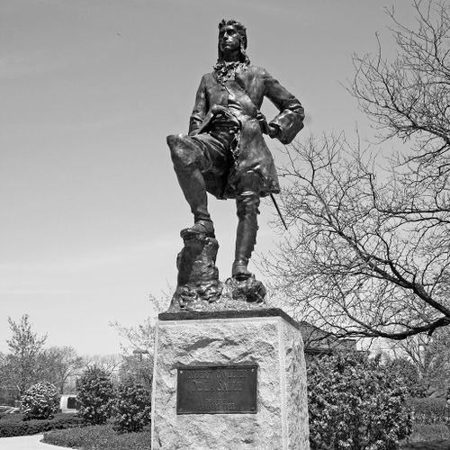 Armed with a pistol and sword and dressed in a rugged frock coat and knee-high, laced boots, the pose of explorer De La Salle is notably swashbuckling.  He stands on a pedestal with his right foot perched on a tree stump and his left hand on his engaged, left hip. 