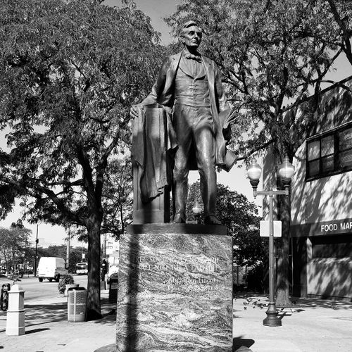 This version of Abraham Lincoln shows the figure standing on a tall, simple pedestal at the corner of Western and Lincoln Avenues near Lincoln Square. Shown on the eve of his presidency, he stands about to speak, next to a draped lectern, and wears his typical long coat, vest and foulard bow tie.