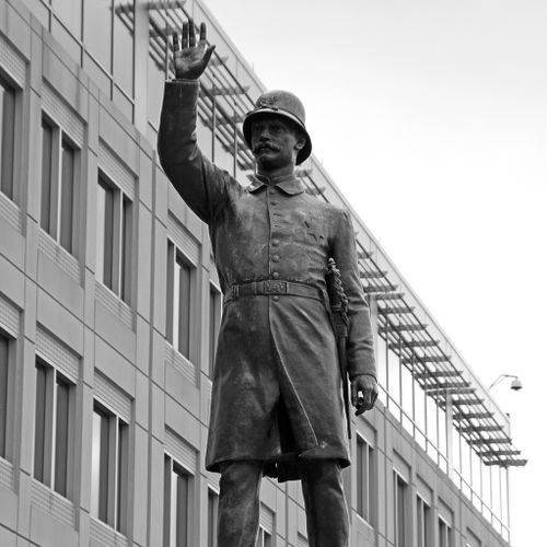 Striding forward slightly atop a high pedestal, a mustachioed figure of a policeman in late 19th-century uniform, complete with badge, helmet and baton, raises his right arm to signal his authority. 