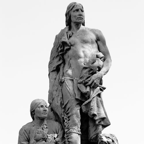 The Alarm is a large scale bronze sculpture atop a tall, limestone pedestal that shows an American Indian family group, including a standing father and a seated mother and child, along with a protective dog. The father, peers off in the distance with an expression of watchful concern.