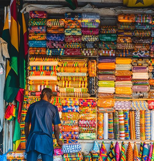 A man with his back towards the camera tending to his shop wall filled with brightly colored woven goods