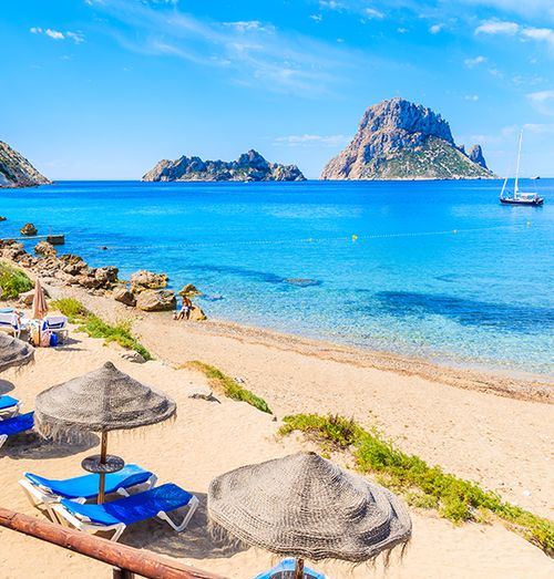 A beautiful beach with blue lounge chairs and tan umbrellas on shore next to crystal blue water with a boat and a couple rocky islands in the distance