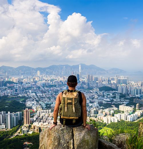 A person with a backpack sitting on a rock and staring out at a view of a large city with mountains in the background on a cloudy day