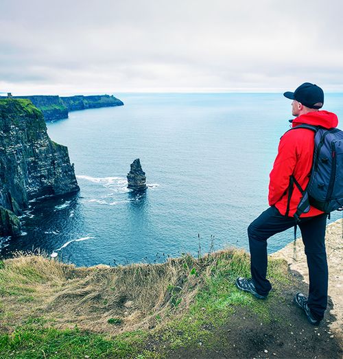 Man in red jacket and black hat with a backpack looks out towards the edge of a cliff next to the ocean on a cloudy day