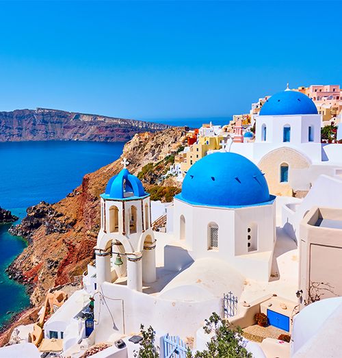 White buildings with bright blue domes on top situated at the edge of a hill next to other brightly colored buildings overlooking the calm sea