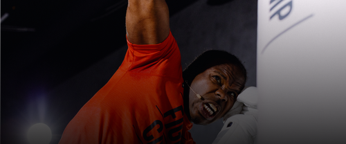 Coach Justin in a red shirt with "FightCamp" printed, wearing a headset microphone, intensely punches a boxing bag indoors.