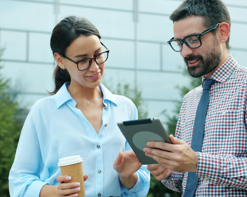 Two professionals with glasses discuss something on a tablet outdoors; one holds a coffee cup, and they're standing in front of a modern building.
