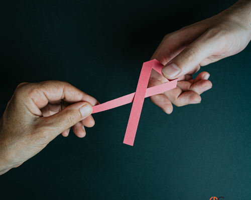 Two hands holding a pink paper ribbon, symbolizing breast cancer awareness, against a dark background.