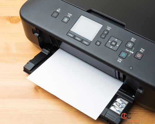 A black inkjet printer with a digital display, showing a blank sheet of paper being printed on a wooden surface.