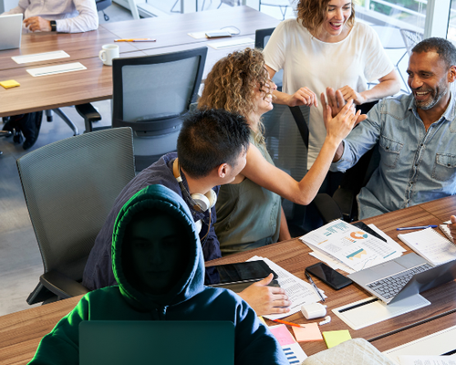 A group of people in an office having a cheerful discussion at a table, while a person in a hoodie works on a laptop in the foreground.