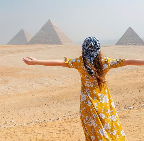 A person with long red hair wearing a yellow floral print dress outstretching their arms and looking at three large pyramid structures located in the desert