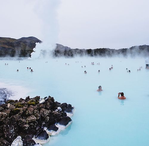 Many people scattered throughout a natural pool filled with pale blue water with steaming rising into the air and black rock formations surrounding the pool