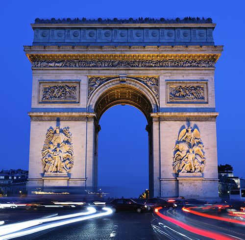 An extremely large beige stone arch with many detailed and symmetrical carvings all over the arch during dusk with long exposure trails of white and red lights at the base of the arch caused by cars driving by