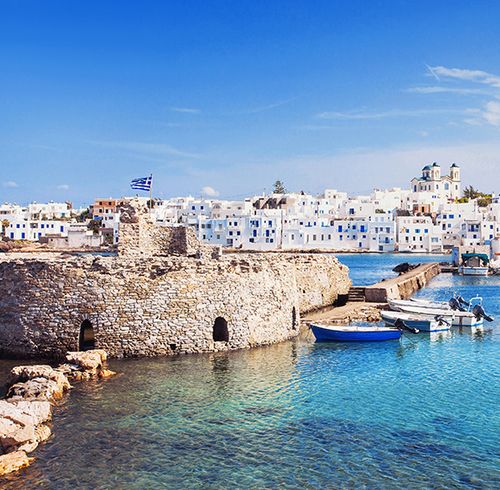 A calm harbor with clear blue water and a stone wall with many densely packed white homes in the background on a clear blue day