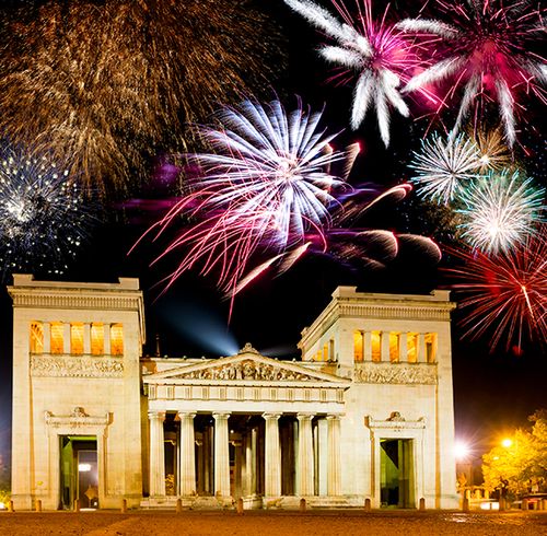 A large grand building with several detailed columns in the front during nighttime with multi-colored fireworks scattered across the sky