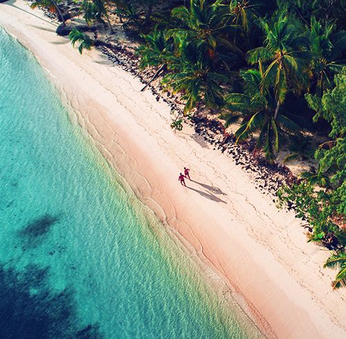 An aerial view of two people walking along the shore of a beach between crystal blue water and a lush green forest
