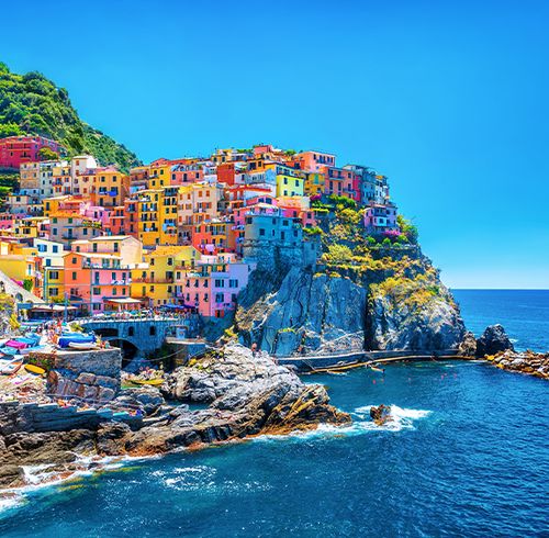 Many brightly colored buildings packed together on a hillside next to an ocean with rocky cliffs with a clear blue sky in the background
