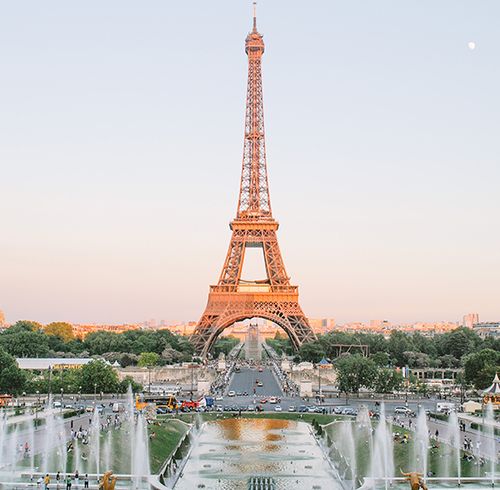 A view of the Eiffel Tower with streets, fountains, cars, people, and a lush forest beneath the tower during dusk