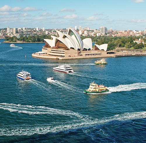 An aerial view of an urban shoreline with large boats traveling through the water and a large white opera house right by the shore with a sprawling city in the background