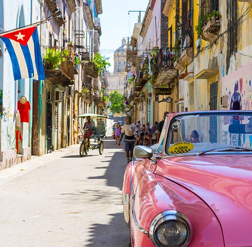A pink vintage car on a city street with a Cuban flag hanging from a blue building