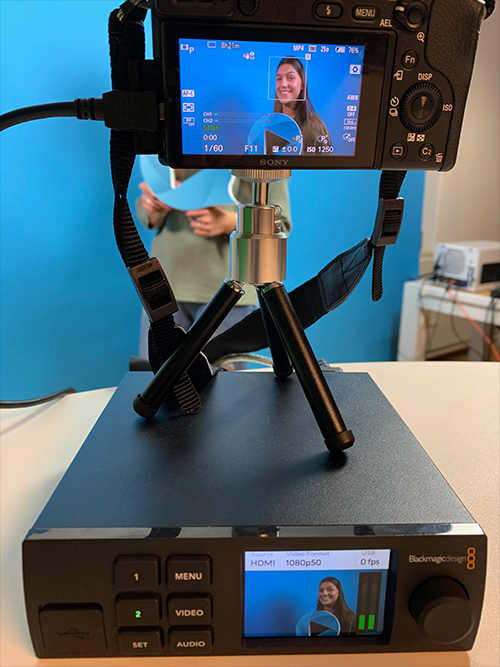 A woman standing behind the camera that's connected to a black box device