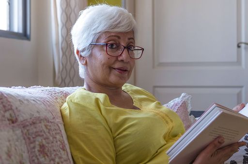Photo of an older lady in respite, looking relaxed, sitting on a lounge holding a book smiling at the camera. She wears a yellow t shirt, glasses and short grey hair