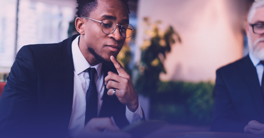 Professional man in glasses thoughtfully reviewing something, a colleague partially visible beside him.