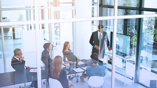 A business meeting in a modern conference room with six people seated at a table, one person standing and presenting, large windows in the background.