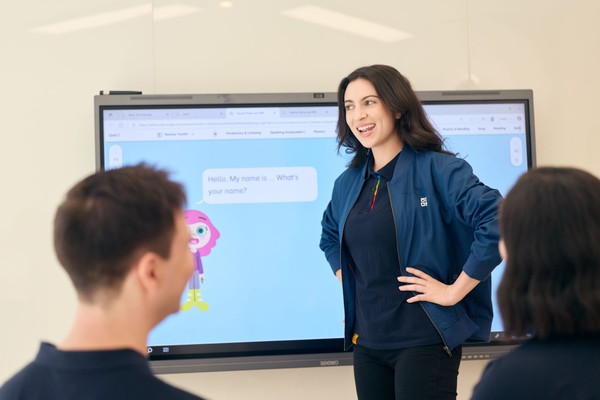 A woman stands smiling in front of a digital screen presenting to two seated individuals. The screen displays a playful character and text bubbles.