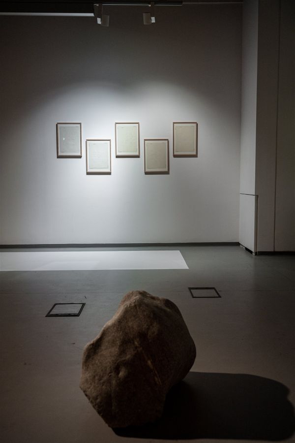 sculpure of a big stone in the front in the back hanging five paper poems in wooden frames | Image by ©