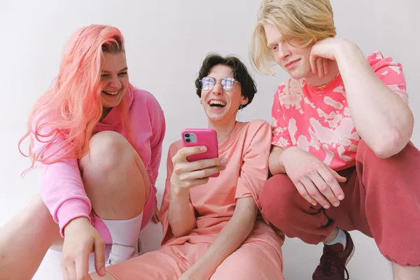 Three people looking at a smartphone together and laughing. Everyone is wearing clothes with pink shades and the phone is also pink. It looks like these youthful people are having a lot of fun.